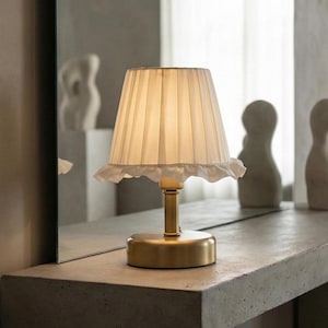 May include: A small, brass-colored table lamp with a pleated white lampshade and a ruffled edge. The lamp has a round base and a slender stem. The lamp is on a concrete surface.
