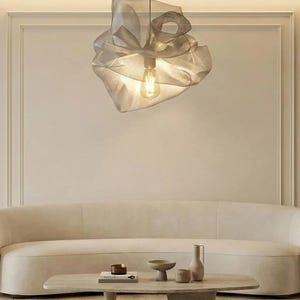 Op de afbeelding: Een moderne, grijze stoffen hanglamp met een decoratief, gerimpeld ontwerp. Het licht hangt aan een dun snoer en verlicht een woonkamer met een neutraal kleurenpalet. De kamer heeft een gebogen bank en een houten salontafel.