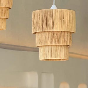 May include: A tiered pendant light fixture with a natural, woven texture. The light has three stacked, cylindrical shades made of light brown material, suspended from a white cord and canopy. The design suggests a bohemian or coastal style.
