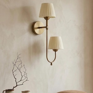 May include: A double-light wall sconce with a brass finish. The sconce features two cream-colored, pleated lampshades. The fixture is mounted on a light-colored wall. Decorative vases and a pillow are on a wooden surface below.