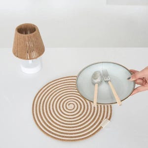 May include: A table setting with a beige and white spiral placemat. A light blue plate with a silver spoon and fork with wooden handles is held above the placemat. A small lamp with a woven shade is also present. The setting is on a white surface.