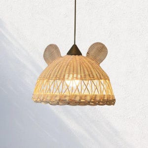 May include: A woven rattan pendant light fixture shaped like a bear's head. The light has two ear-shaped accents and a conical top. The light is suspended from a dark cord and is lit from within, casting a warm glow.