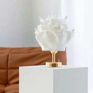 May include: A white, rose-shaped table lamp with a gold base. The lamp's shade is made of layered, petal-like material. The lamp sits on a white cube, with a brown leather sofa in the background. The lamp is a decorative lighting fixture.