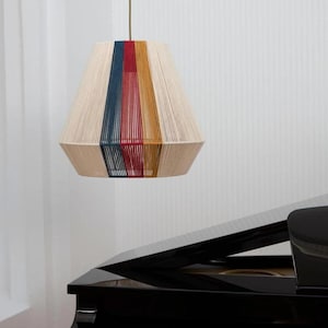 May include: A beige pendant light with vertical stripes of blue, red, and yellow. The lampshade is a trapezoid shape, suspended by a thin, brown cord. The background is a white wall, and a black piano is visible in the foreground.