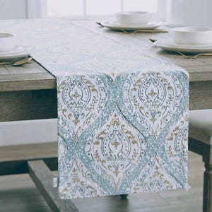 May include: A rectangular table runner featuring a repeating damask pattern in shades of light blue, white, and muted gold. The fabric displays a distressed, weathered texture throughout the intricate floral and scrollwork design elements.