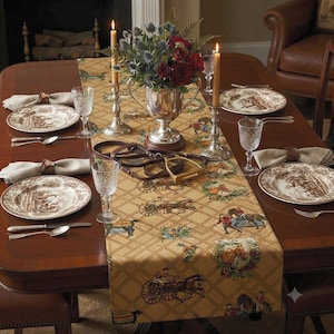May include: A tan table runner featuring a repeating equestrian pattern of horses, carriages, and riders in a diamond grid. The runner is set on a wooden table with white plates, crystal glasses, silver candlesticks, and a floral centerpiece.
