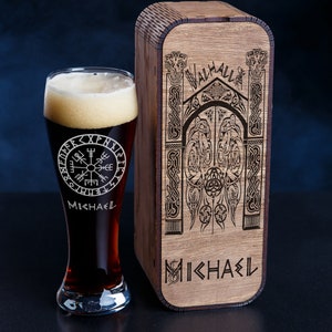 May include: A wooden box with a Viking-themed design and the word "Valhalla" engraved on it. The box is closed and has a dark brown finish. The box is sitting on a black surface next to a clear glass beer stein with a black liquid inside. The stein has a compass design and the name "Michael" engraved on it.