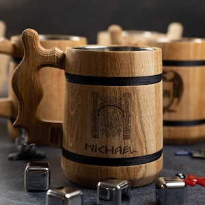 May include: Wooden beer tankards with decorative handles and stainless steel interiors. The tankards have a light brown finish with black accents and are engraved with a gate design and the name "MICHAEL". Several metal ice cubes are in front of the tankards.