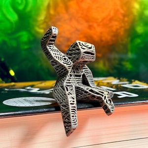 May include: A small, metallic-colored figurine with a complex, lattice-like geometric structure. The figure has a boxy head, a torso, and limbs, all featuring an intricate, interconnected web pattern throughout its entire surface.
