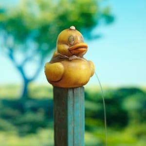 May include: A yellow rubber duck with painted eyes and an orange beak, featuring a thin, clear plastic string tied around its neck. The duck is mounted on top of a weathered, rectangular, teal-colored post with visible signs of wear and texture.