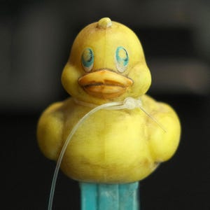 Könnte beinhalten: Eine gelbe Quietscheente mit blauen Augen und einem orangefarbenen Schnabel, um deren Hals ein durchsichtiger Kunststoff-Kabelbinder gebunden ist, montiert auf einem hellblauen rechteckigen Sockel. Die Ente hat eine verwitterte, texturierte Oberfläche.
