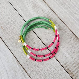 Puede incluir: Una pulsera envolvente de cuentas con tres hilos. El hilo superior es verde claro, el hilo del medio es rosa con cuentas negras y el hilo inferior también es verde claro. La pulsera tiene dijes dorados en forma de corazón.