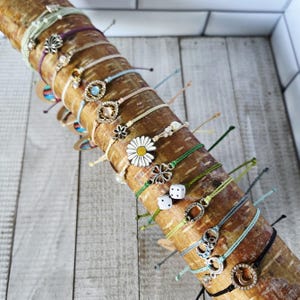 May include: A collection of colorful friendship bracelets with various charms, including a daisy, a four-leaf clover, dice, and a horseshoe. The bracelets are displayed on a wooden log.