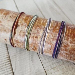 May include: Five colorful string bracelets are displayed on a wooden log. The bracelets feature various color combinations, including red, green, white, and purple. The log has a natural, textured appearance, and the background is a light wood surface.