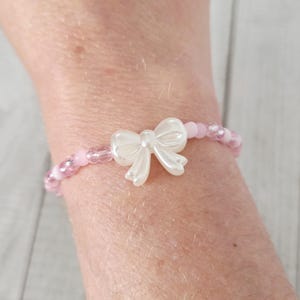 Puede incluir: Una pulsera de cuentas rosas con un charm de lazo blanco. La pulsera presenta pequeñas cuentas rosas redondas y un lazo blanco nacarado en el centro. Un accesorio encantador.