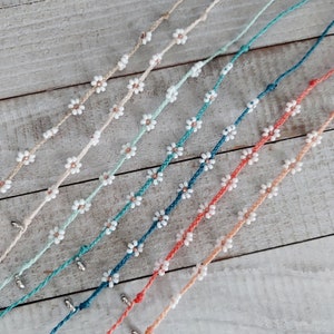 May include: A set of seven colorful bracelets with white daisy flower beads. The bracelets are made with different colored cords and are arranged in a fan shape.