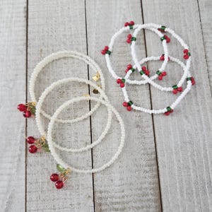 Op de afbeelding: Verschillende armbanden met kralen. Sommige zijn wit met rode kersenbedels en groene blaadjes. Andere zijn wit met rode en groene kralen. De armbanden liggen op een licht houten oppervlak.