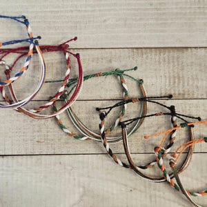 Op de afbeelding: Een verzameling kleurrijke gevlochten armbanden met verschillende kleurencombinaties, waaronder blauw, rood, wit, groen en oranje. De armbanden liggen op een lichtgekleurd houten oppervlak.