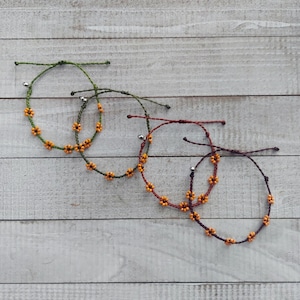 May include: Three beaded bracelets with orange flower charms and silver bell accents. The bracelets are made with green, brown, and dark brown cord.