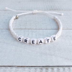 May include: White macrame bracelet with the word "CREATE" spelled out in black letters on white cube beads. The bracelet has a sliding knot closure and a small silver bead.