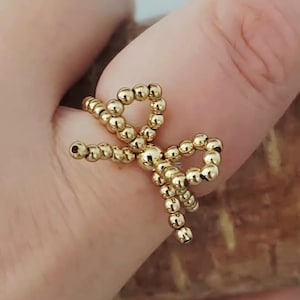 May include: A gold-colored bow ring made of small, round beads. The ring is a delicate piece of jewelry, with the bow shape clearly defined. The beads catch the light, creating a subtle sparkle. The ring is shown on a finger.