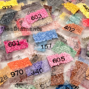 May include: Assortment of small plastic bags filled with colourful diamond-shaped beads, labelled with numbers. The bags are arranged in a random pattern on a white surface.