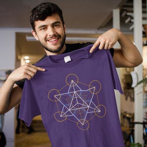 Könnte beinhalten: Ein lila T-Shirt mit einem geometrischen Design aus goldenen, weißen und blauen Linien. Das Design umfasst Kreise, Dreiecke und andere geometrische Formen. Das T-Shirt ist einfarbig und hat einen Rundhalsausschnitt.
