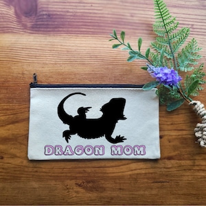 May include: A white canvas zippered pouch with a black silhouette of a bearded dragon and the text "Dragon Mom" in pink.