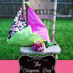May include: A green, pink, and black fabric origami bag with a floral pattern sits on a wooden chair. The bag is folded into a triangular shape and has a handle. The chair is in a grassy area with a pink banner that reads "The Origami Bag".