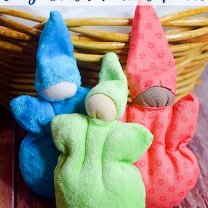 May include: Three small cloth pocket dolls in a wicker basket. The dolls are made of soft fabric and have simple features. The dolls are blue, green, and red.