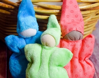 Può includere: Tre piccole bambole di stoffa da tasca in un cesto di vimini. Le bambole sono fatte di tessuto morbido e hanno tratti semplici. Le bambole sono blu, verdi e rosse.