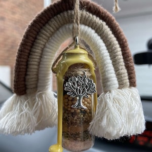 May include: A macrame rainbow in shades of brown, beige, and white, with a glass bottle pendant. The bottle contains various dried herbs and seeds, sealed with yellow wax and a silver tree charm. The pendant hangs from a wooden bead.