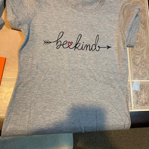 Puede incluir: Camiseta gris con una flecha negra y el texto "be kind" con un corazón rosa en el medio.