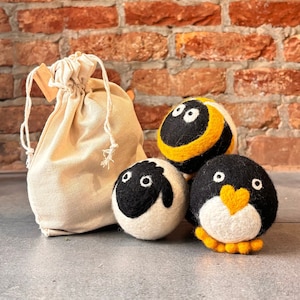 May include: A set of three felted wool animal-shaped balls: a sheep, a bee, and a penguin, with a drawstring bag. The sheep is white with a black face, the bee is black and yellow, and the penguin is black and white with a yellow heart-shaped beak.