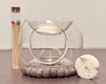 May include: A glass wax melter with a round base and a removable bowl. The melter has a small tea light holder in the centre. The melter is sitting on a grey felt coaster. A small white wax melt is next to the melter.