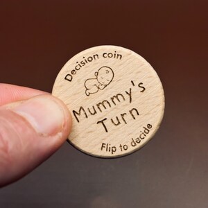 以下が含まれることがあります： 赤ちゃんが描かれた小さなイラストと「Decision coin Mummy's Turn Flip to decide」という文字が書かれた木製の硬貨です。