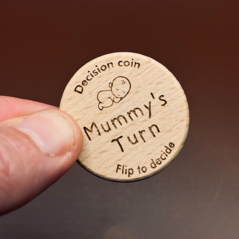 以下が含まれることがあります： 赤ちゃんが描かれた小さなイラストと「Decision coin Mummy's Turn Flip to decide」という文字が書かれた木製の硬貨です。