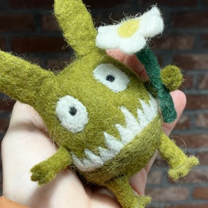 May include: A green felt monster with white teeth and eyes, holding a white felt flower with a yellow center. The monster has a green felt stem and is holding the flower in its right hand.