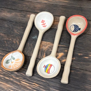 Könnte beinhalten: Vier Keramiklöffel mit Holzgriffen. Jeder Löffel hat ein anderes Design: eine Blume, ein Fisch, eine Pflanze und ein gestreifter Fisch. Die Löffel liegen auf einer Holzoberfläche.