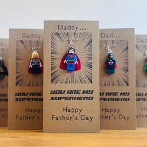 May include: Five Father's Day cards with superhero Lego figures attached. Each card has the text "You Are My Superhero" and "Happy Father's Day". The cards feature Batman, Thor, Superman, a black and red superhero, and a green superhero.
