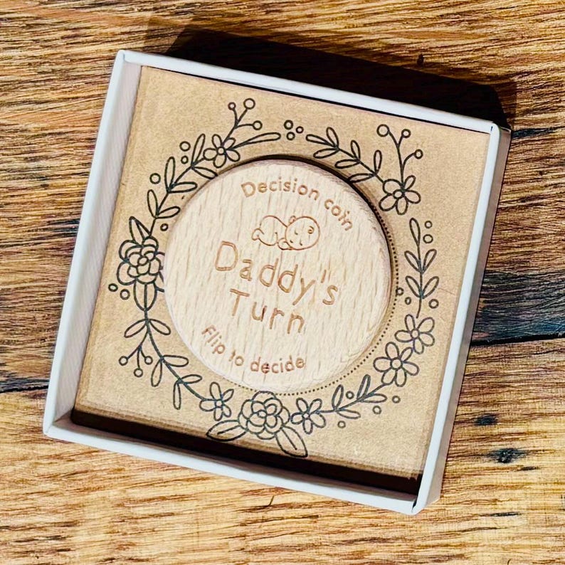 以下が含まれることがあります： 縁に花柄が施された木製の硬貨。硬貨には「Decision coin」と「Daddy's Turn」の文字が書かれている。硬貨には、寝ている赤ちゃんの小さなイラストも描かれている。「Flip to decide」という文字が硬貨の下部に印刷されている。