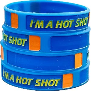 Könnte beinhalten: Drei blaue Silikon-Armbänder, die übereinander gestapelt sind. Auf jedem Band steht in gelben Buchstaben "I'M A HOT SHOT". Auf jedem Band sind auch orangefarbene rechteckige Akzente zu sehen.