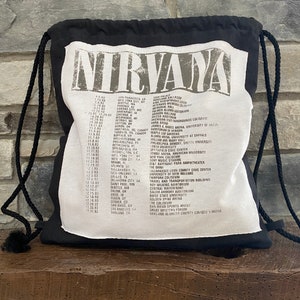 May include: Black drawstring bag with a white graphic featuring the band name "NIRVANA" and a list of tour dates and locations. The bag is made of a soft material and has black drawstrings.