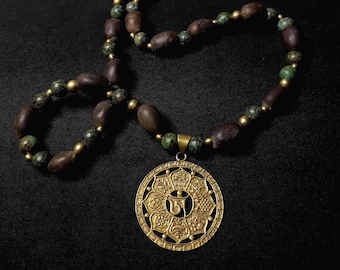 Könnte beinhalten: Halskette mit einem verzierten, goldfarbenen Anhänger mit einem zentralen Design und filigranen Details. Die Kette hat eine Perlenkette mit braunen und grünen Perlen und goldfarbenen Abstandshaltern. Der Hintergrund ist schwarz.
