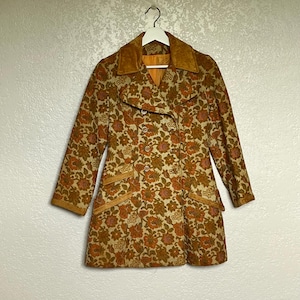 May include: A double-breasted coat featuring a tan and orange floral pattern throughout. The garment includes a solid tan collar, matching tan pocket flaps, and a series of buttons down the front. The coat is displayed on a white hanger.