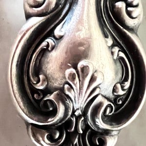 Könnte beinhalten: Ein silberfarbener Löffelstiel aus Metall mit einem komplizierten geprägten Schnörkelmuster. Das Design umfasst symmetrische geschwungene Linien und ein zentrales fächerförmiges Motiv an der Basis, mit einer polierten Oberfläche.