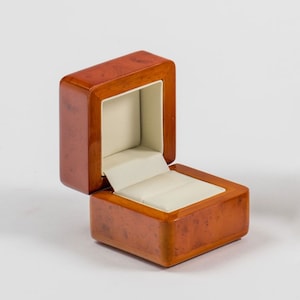 May include: A wooden ring box with a polished finish. The box is open, showing a cream-coloured interior. The box is rectangular with rounded edges and a hinged lid. The wood grain is visible.