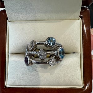 May include: A silver ring with multiple bands and a variety of blue and clear gemstones. The ring is in a white velvet ring box.