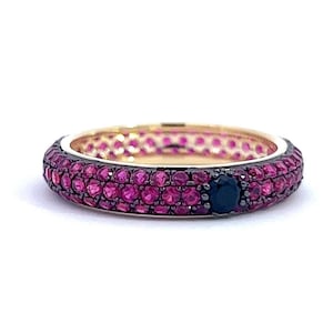 May include: A gold ring featuring a band encrusted with small, round, pink gemstones. A single, dark blue gemstone is set into the band. The ring is set against a white background.