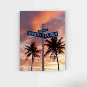 May include: A sunset scene with two palm trees silhouetted against the sky. A street sign with the names "Carlos" and "Lauren" is mounted on a pole. The sign reads "Bridge 2024".
